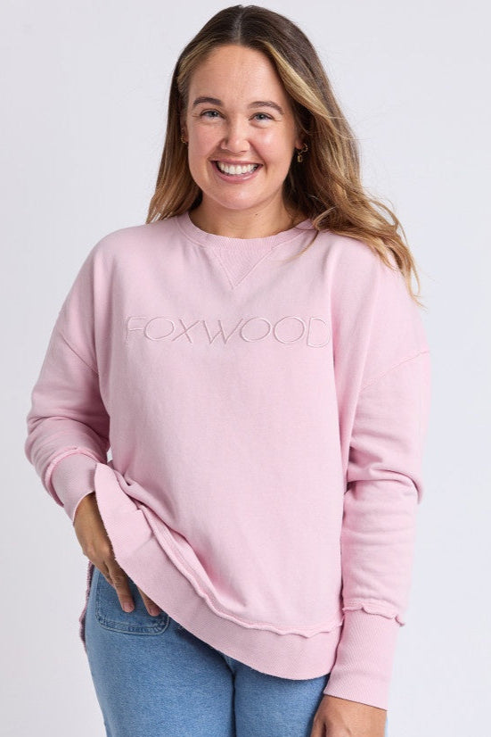 A woman smiling and wearing a pink sweatshirt with the brand name 'Foxwood' on the front, paired with blue jeans.