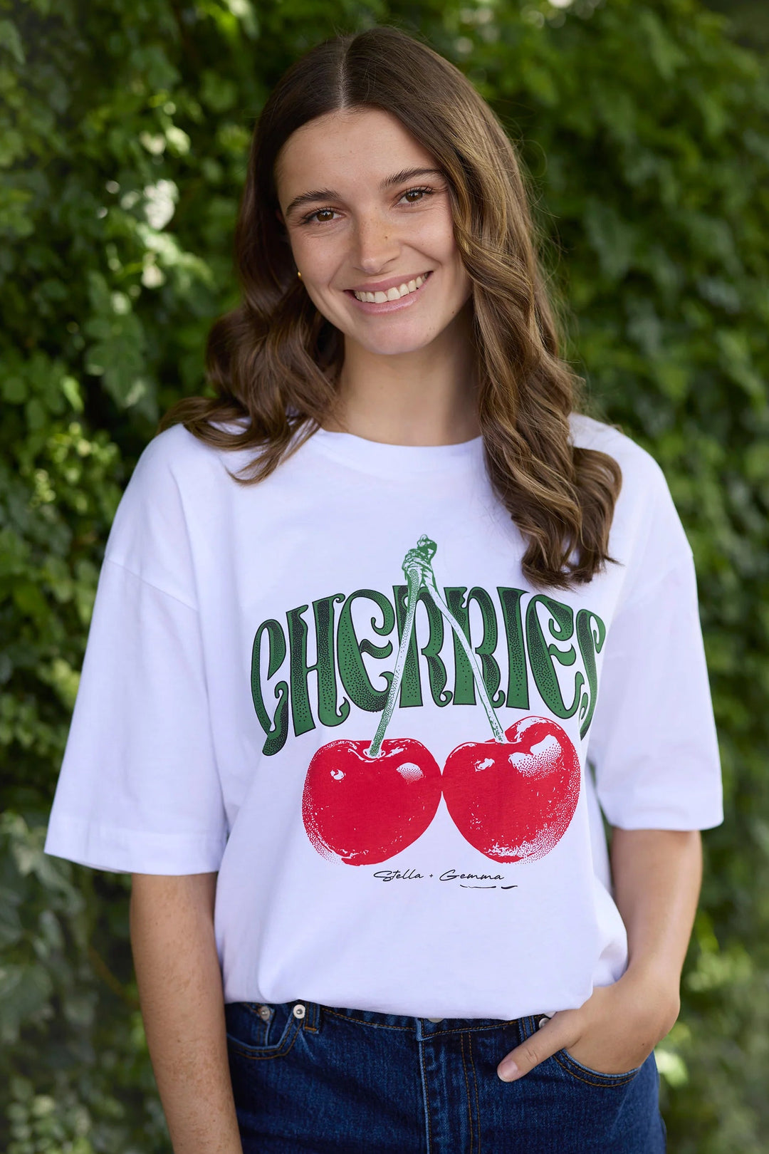 Person wearing a white t-shirt with red cherries and green text, standing against a green leafy background.