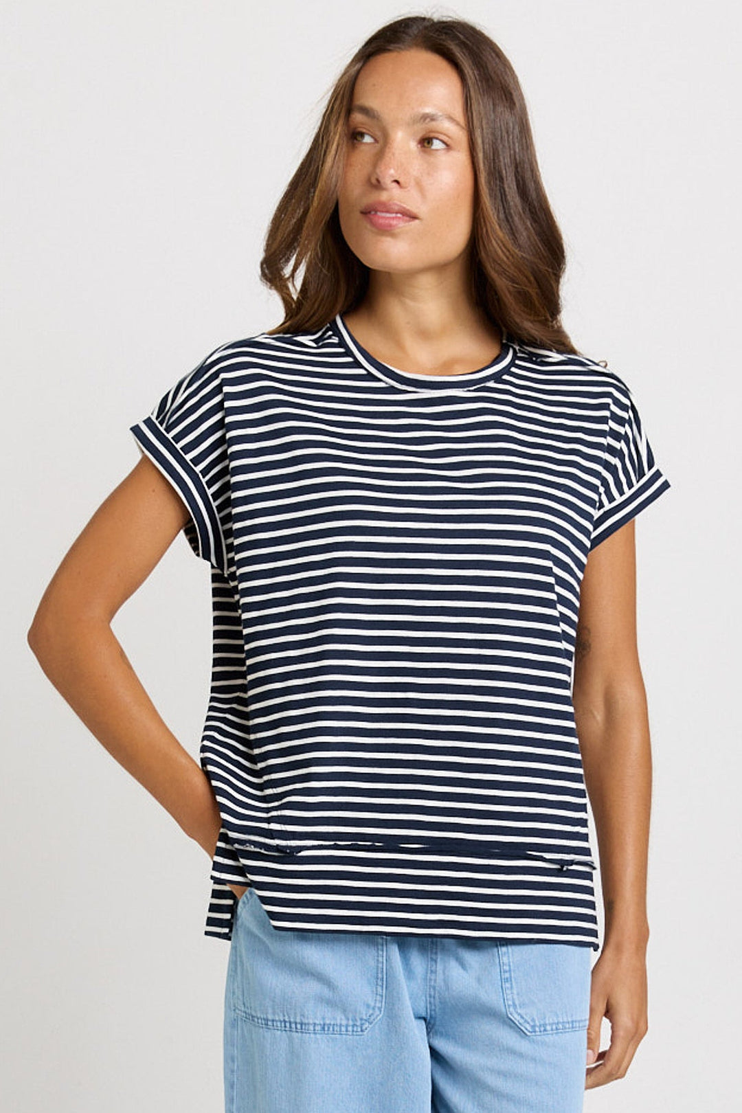 A woman wearing a navy and white horizontally striped short-sleeve t-shirt with light blue jeans, standing against a light gray studio background.