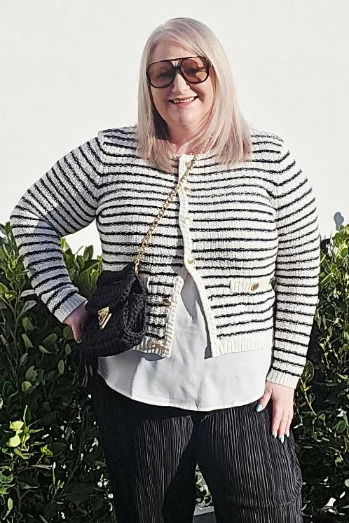 A woman wearing a black and white striped cardigan over a white top and black pinstripe trousers, standing outdoors on a concrete path in front of a white wall and green bushes.