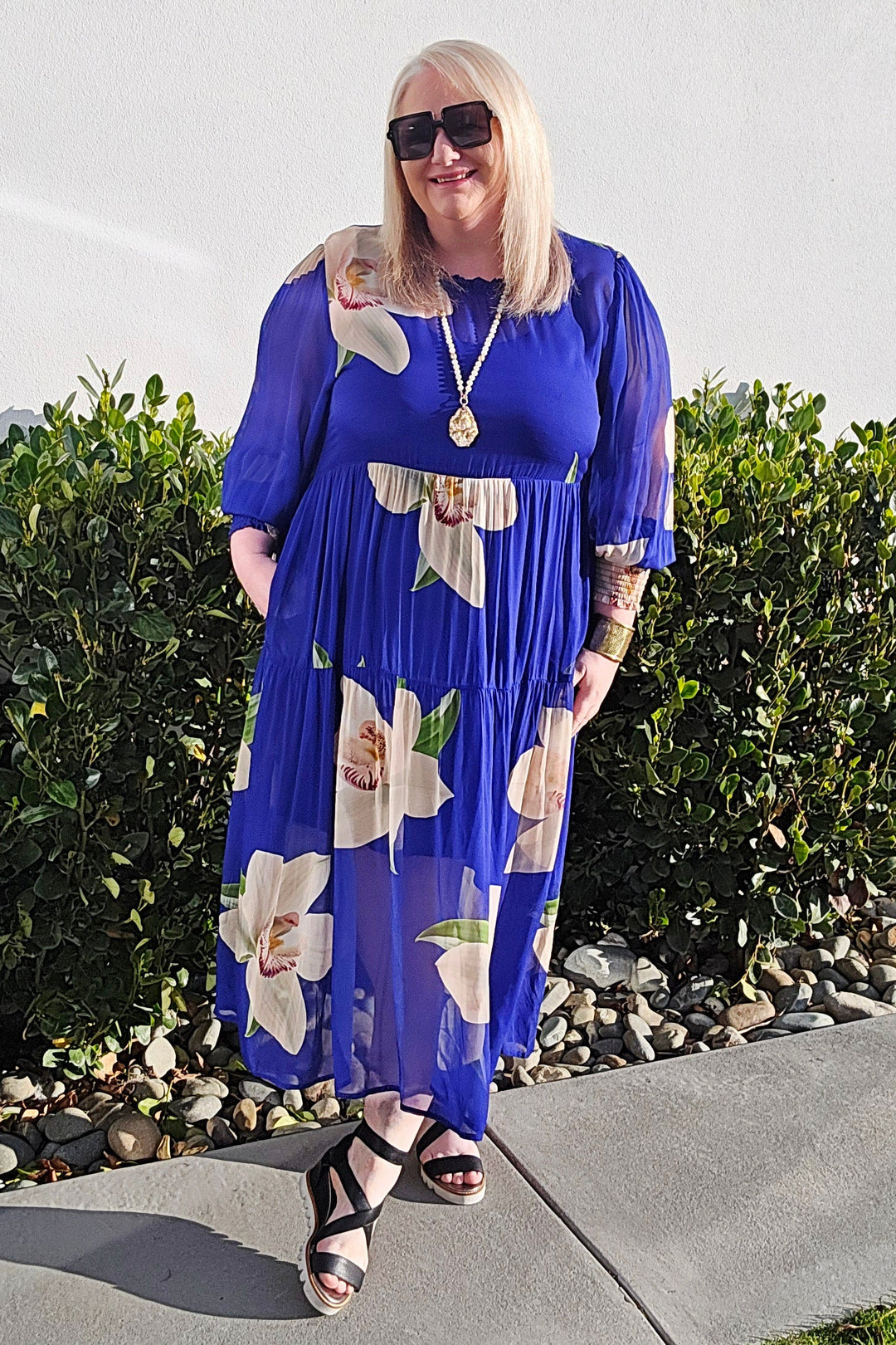 A woman wearing a royal blue midi dress with a white floral print, standing outdoors on a concrete walkway in front of a white wall and green bushes.