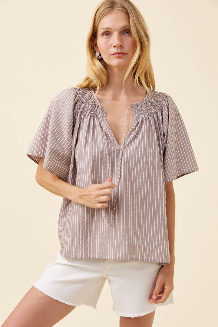 A woman modeling a short-sleeve smocked blouse with vertical stripes in a muted taupe-brown and beige color, paired with white shorts, against a solid cream background.