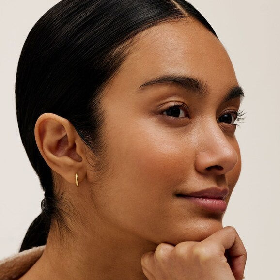 Woman with a neutral expression wearing a gold TED BAKER earring against a beige background