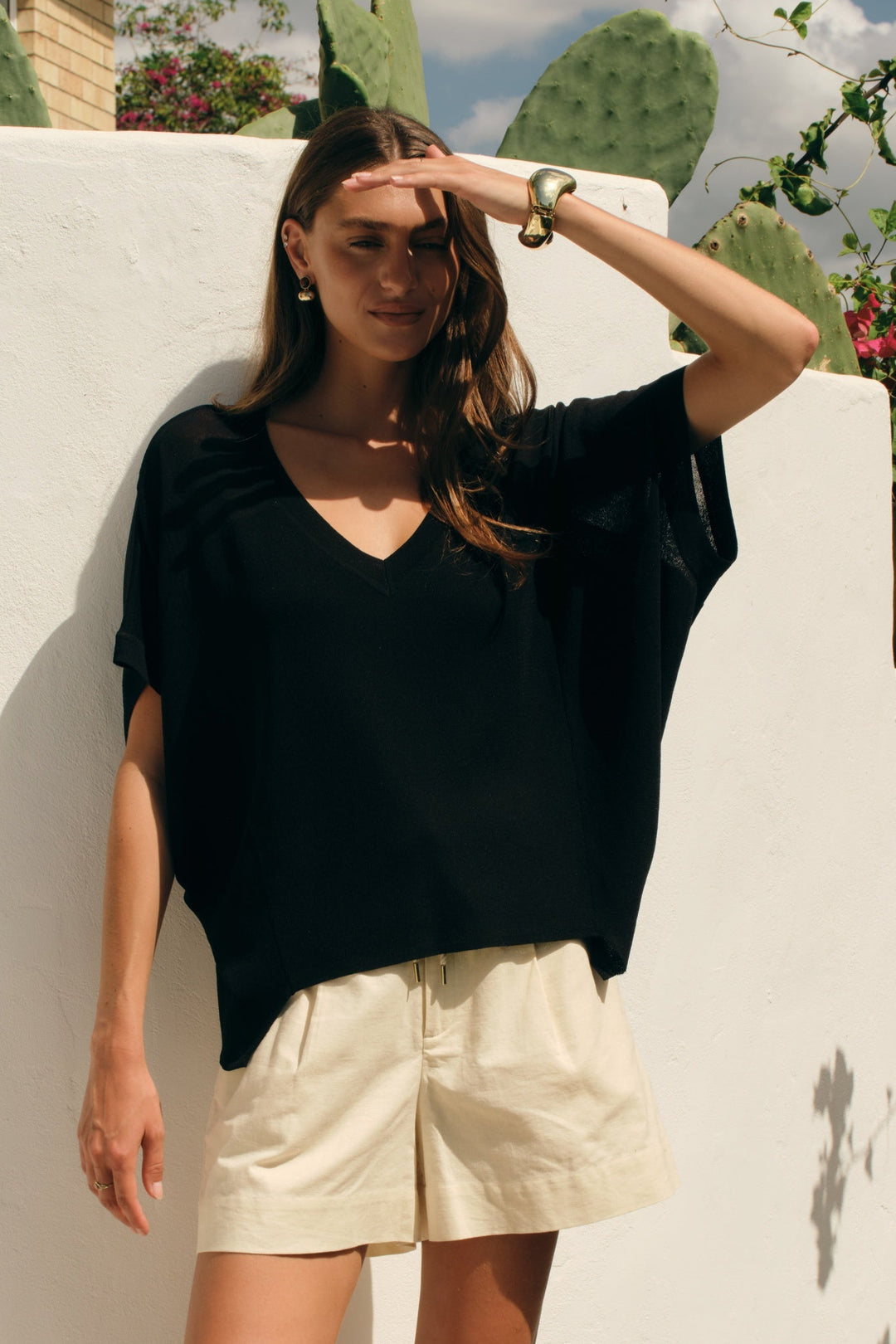 A woman stands outdoors against a white wall wearing a loose black short-sleeve top and light beige shorts, with cacti, flowers, and a house visible in the background.