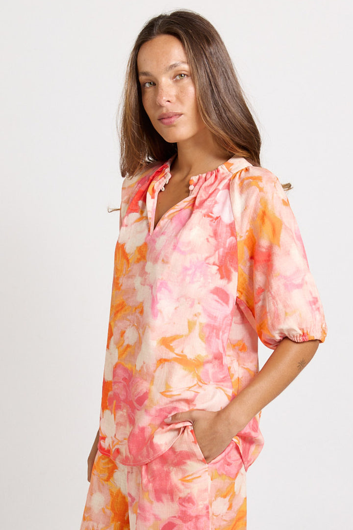 A woman modeling a short-sleeve, loose-fitting blouse with a pink, orange, and white watercolor floral print, set against a plain light gray background.