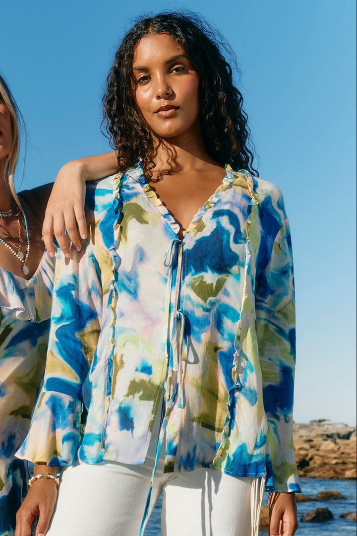 Two women standing on a sandy beach wearing matching blue, green, and white abstract print clothing, with the ocean and blue sky in the background.