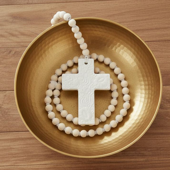 A white embossed resin cross ornament with floral and scroll patterns, hanging from a cream-colored wooden bead chain against a plain white background.