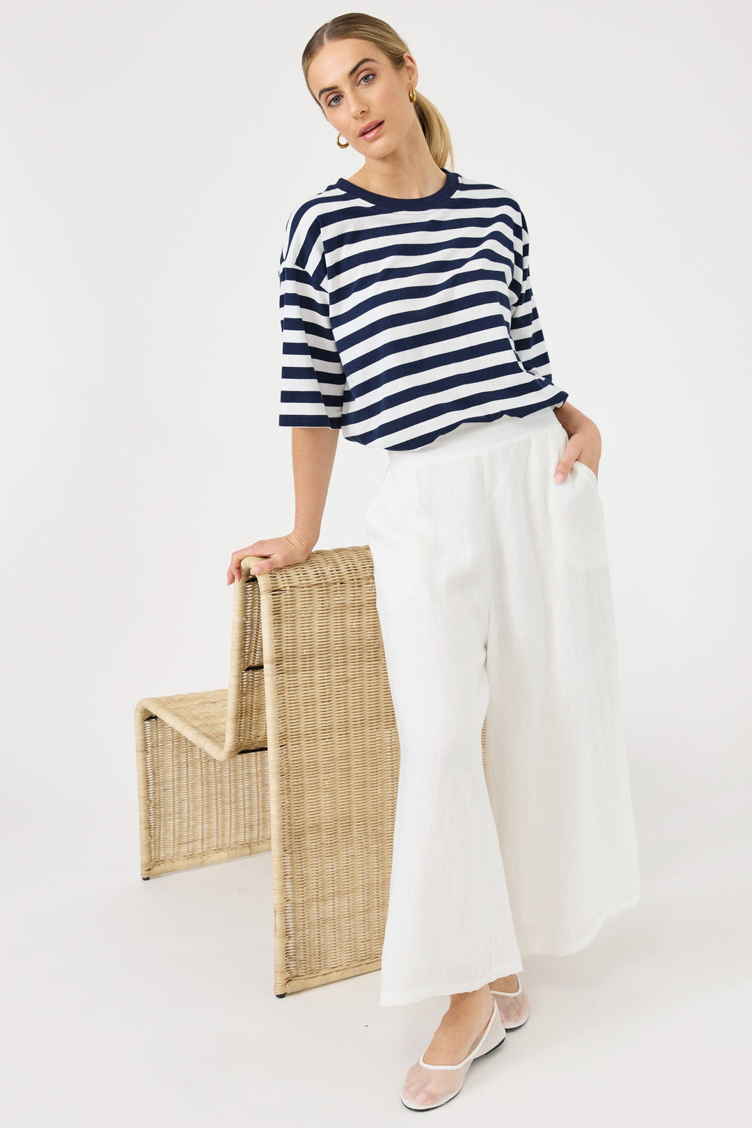 A woman modeling a navy and white striped short-sleeve top with white wide-leg pants, standing next to a woven wicker chair against a plain white studio background.