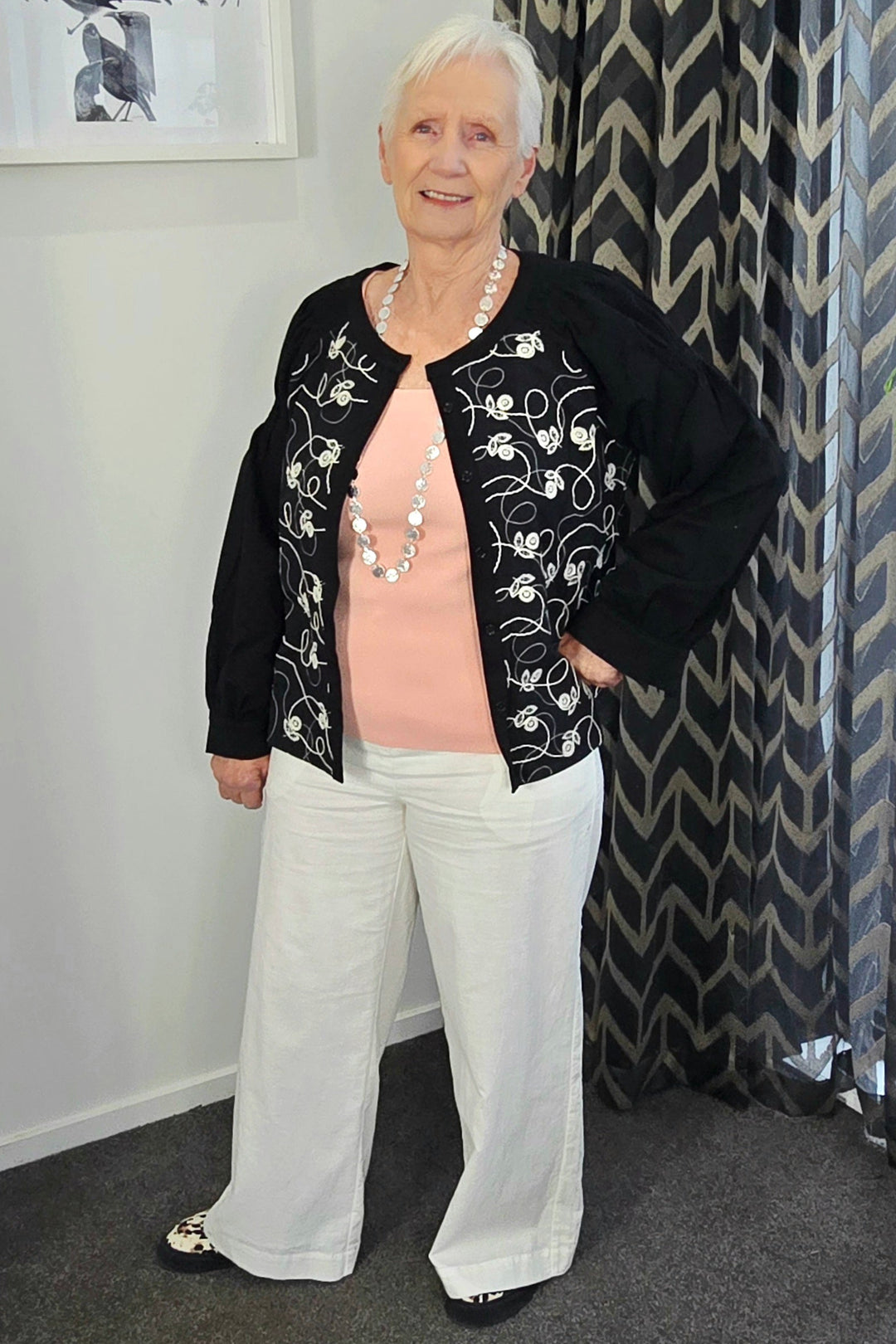 Woman wearing a black embroidered jacket and white pants standing in a room with a patterned curtain.