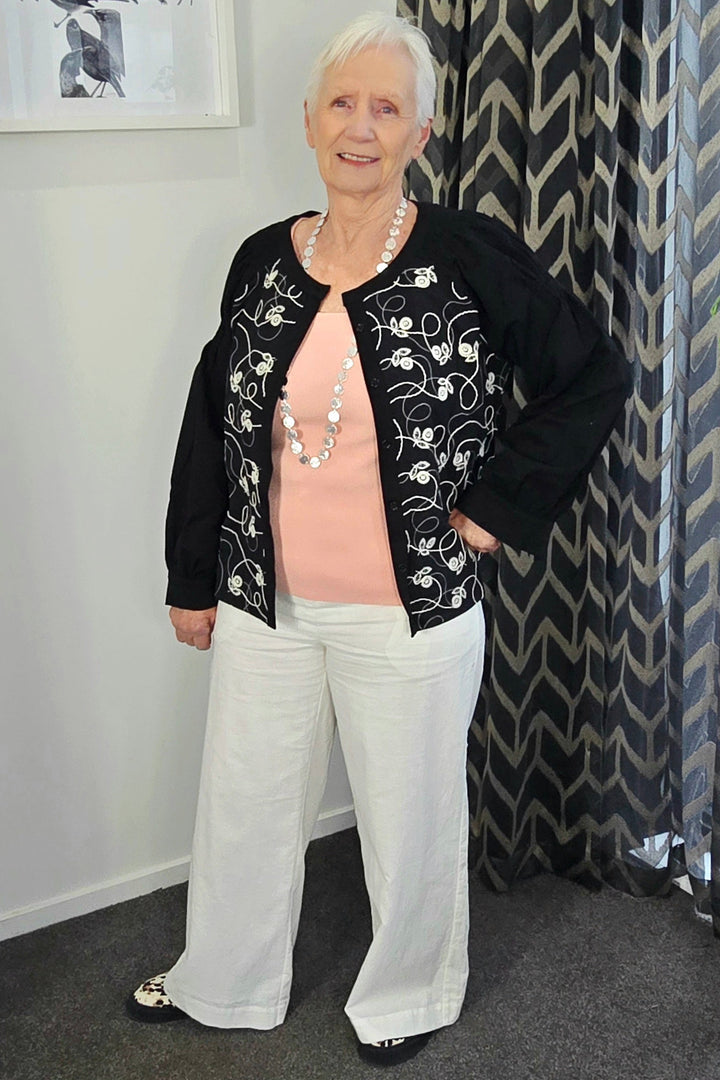Woman wearing a black embroidered jacket and white pants standing in a room with a patterned curtain.
