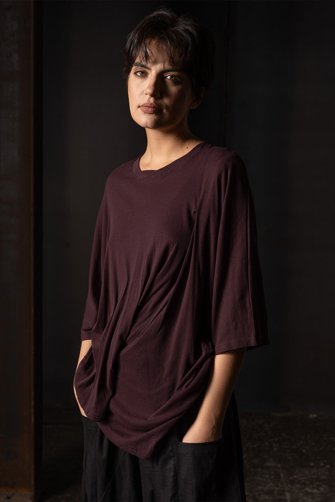 A woman wearing a loose, dark plum-colored tunic top with draped details and black pants, standing in a dark, moody indoor setting.