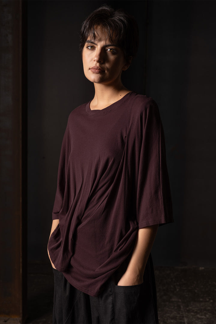 A woman wearing a loose, dark plum-colored tunic top with draped details and black pants, standing in a dark, moody indoor setting.