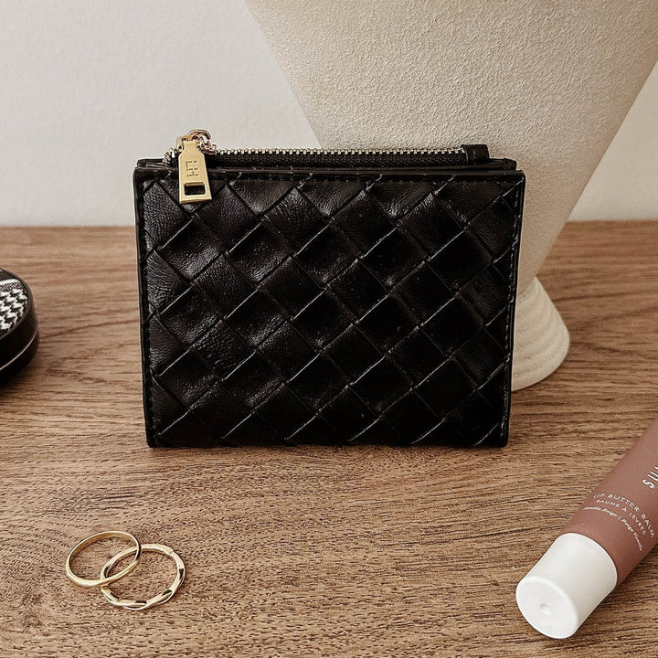 Black quilted louenhide wallet, gold rings, and a brown tube on a wooden surface with a white vase in the background.