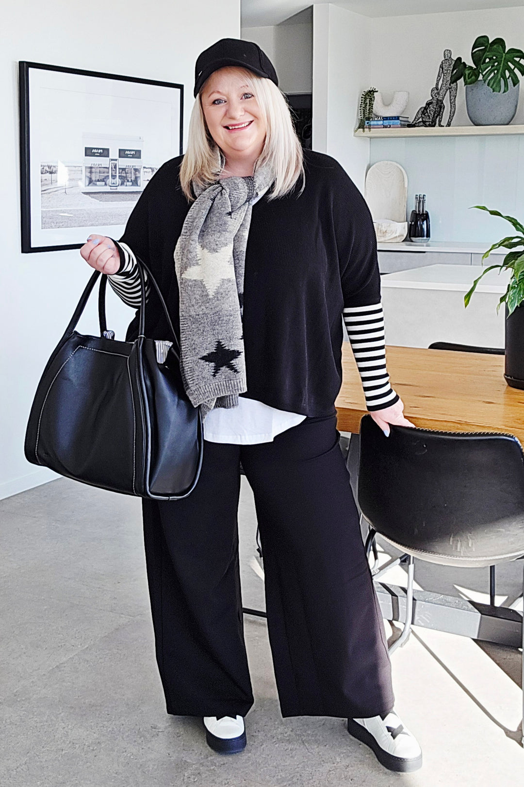 Woman in a stylish outfit with a black sweater, scarf, and pants, holding a black handbag in a modern room.