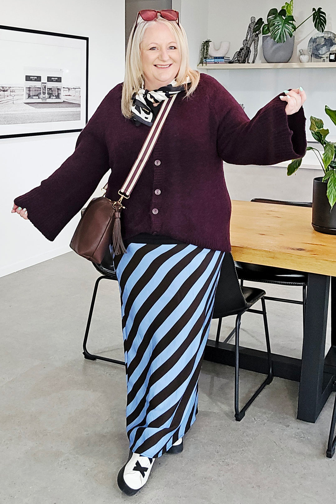 A woman modeling a dark purple knit cardigan sweater paired with a black and light blue striped maxi skirt, a brown shoulder bag, and white sneakers with black bows, set in a modern indoor room with a wooden table and plants.
